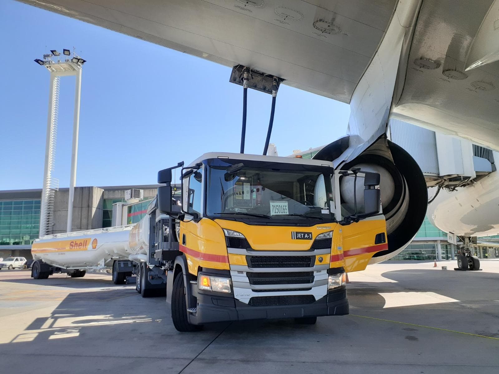 A Shell Jet A-1 fuel truck refuels an aircraft on an airport apron, with its hoses connected under the wing.