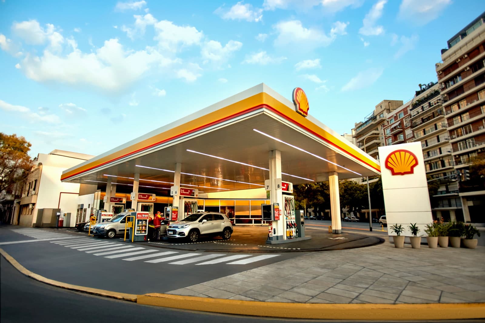 A Shell service station in daytime, with cars refueling under the canopy and apartment buildings behind.