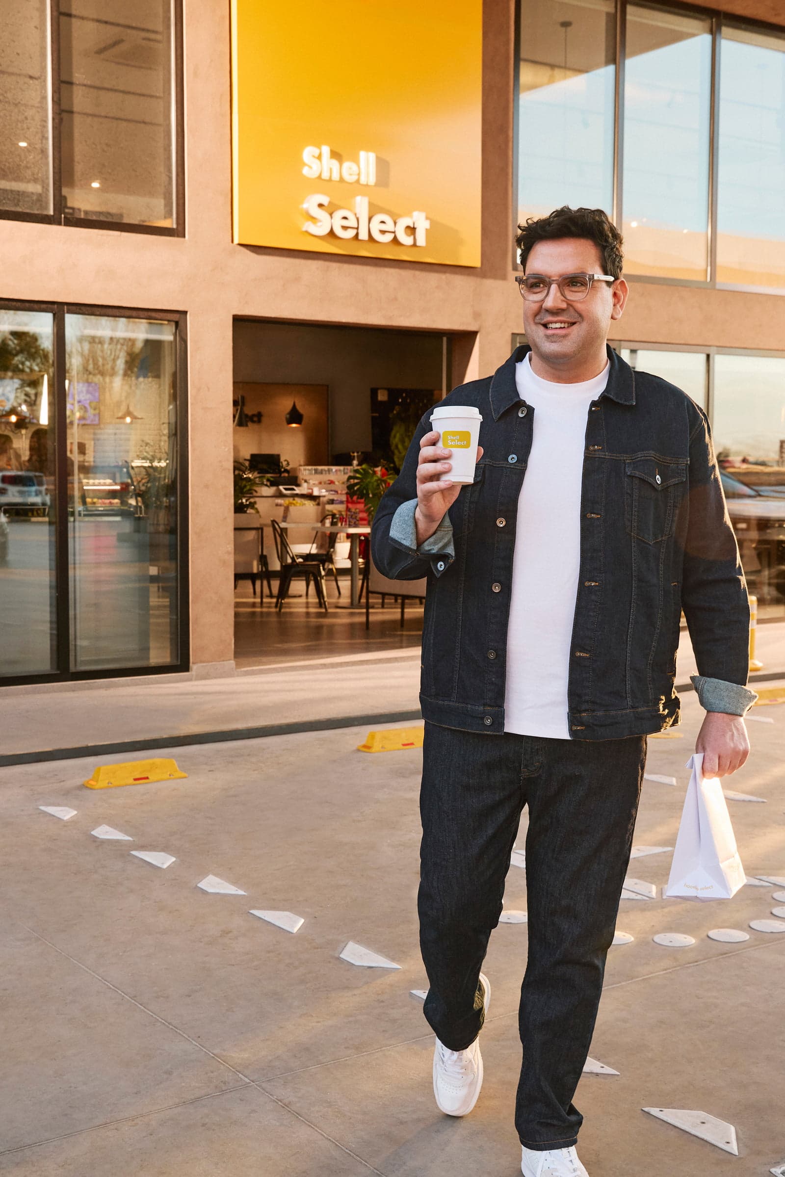 A smiling man walks out of a Shell Select store with a coffee and a shopping bag.