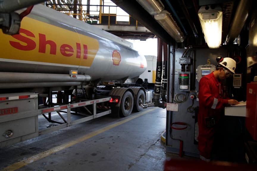 A worker in a hard hat and workwear checks the loading of a tanker truck at a dispatch terminal.